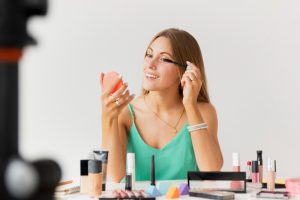 Mulher sorridente aplicando rímel enquanto se olha em um espelho compacto, com diversos produtos de maquiagem à sua frente.