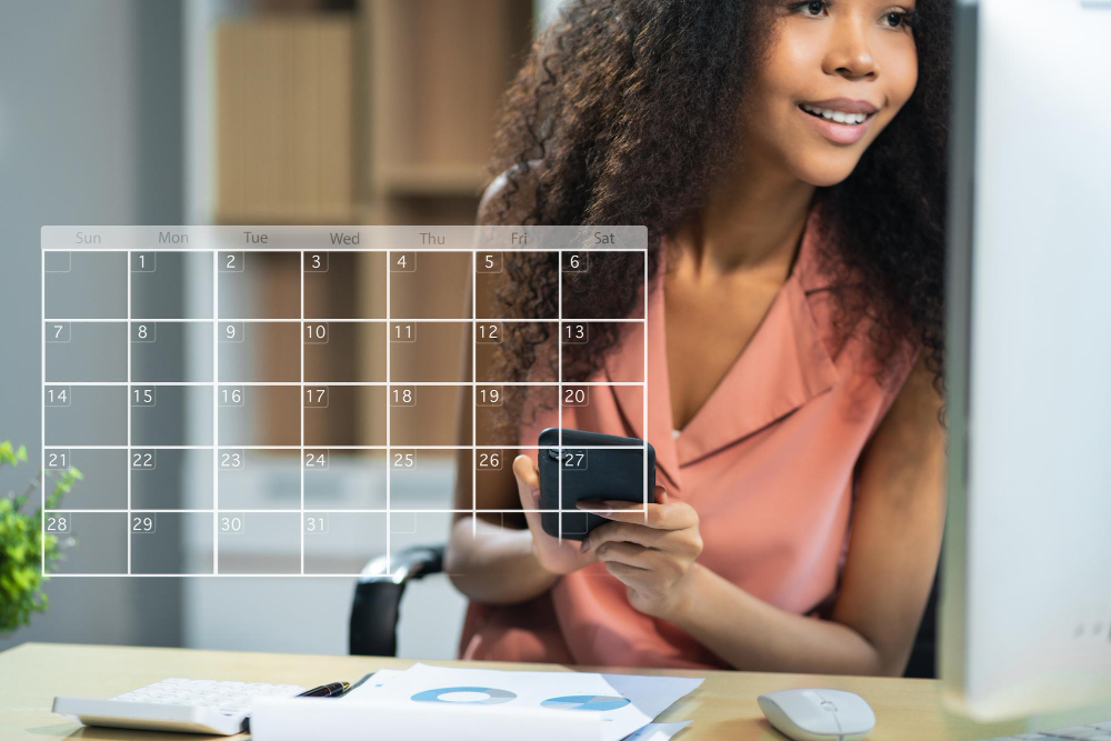 Mulher sorridente com um smartphone na mão, sentada em frente ao computador com uma sobreposição gráfica de calendário.