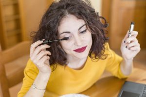 Mulher de cabelo castanho curto aplicando rímel nos cílios enquanto sorri, vestindo uma blusa amarela e sentada em um ambiente interno com móveis de madeira.