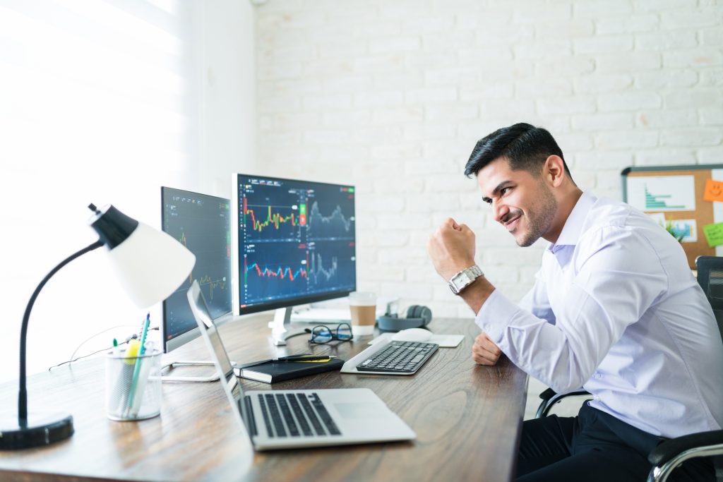 Homem sorridente, vestindo uma camisa social, fazendo um gesto de vitória enquanto observa gráficos financeiros em múltiplos monitores. O ambiente é um escritório com mesas, laptops e objetos de escritório, indicando um trabalho focado em trading ou análise de mercado.