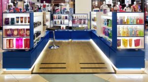 Quiosque moderno em shopping center, com prateleiras iluminadas exibindo diversos produtos de perfumaria e cosméticos. Há um banco alto no centro do espaço, que tem piso de madeira clara e iluminação LED na base das estruturas.