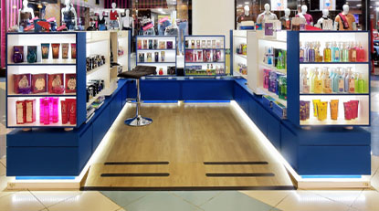 Quiosque moderno em shopping center, com prateleiras iluminadas exibindo diversos produtos de perfumaria e cosméticos. Há um banco alto no centro do espaço, que tem piso de madeira clara e iluminação LED na base das estruturas.