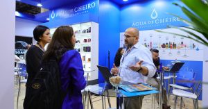 Homem conversa com duas mulheres em estande da marca Água de Cheiro durante uma feira ou evento empresarial.