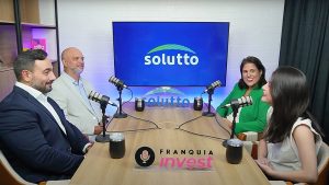 Quatro pessoas gravam um podcast em estúdio com logotipo “Solutto” ao fundo e placa “Franquia Invest” sobre a mesa, conversando de forma descontraída.