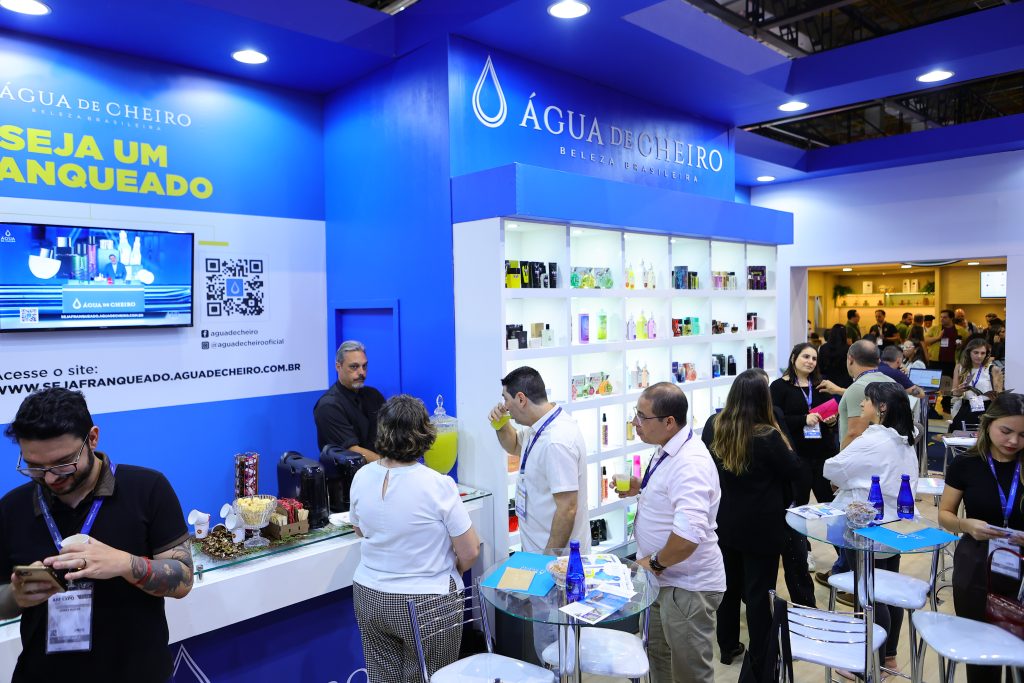 Estande da Água de Cheiro em evento, com visitantes interagindo e produtos expostos em prateleiras iluminadas.