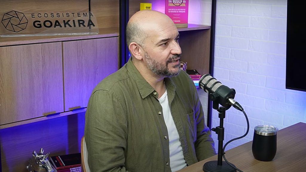 Homem careca com barba participa de podcast, falando ao microfone em estúdio com fundo decorado e placa “Ecossistema Goakira”.
