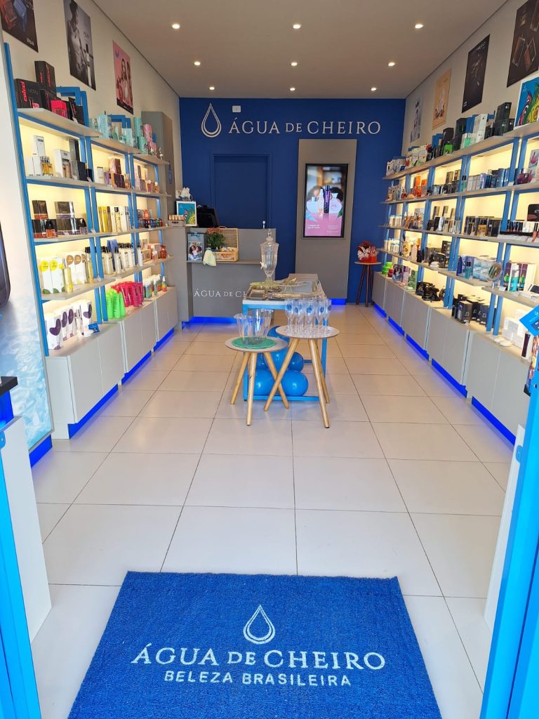 Interior da loja Água de Cheiro, com prateleiras iluminadas repletas de perfumes e cosméticos, tapete azul com a logomarca e mesa central com taças e brindes.