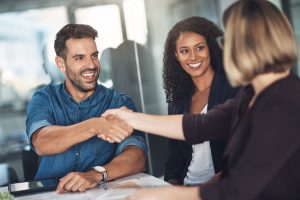 Duas pessoas apertando as mãos em um ambiente corporativo, com outra pessoa sorrindo ao lado, simbolizando um acordo ou parceria.