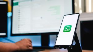 Smartphone com o ícone do WhatsApp na tela sobre suporte, em frente a um computador com o WhatsApp Web aberto.