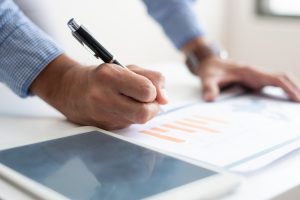Pessoa analisando gráfico impresso enquanto faz anotações com caneta, ao lado de um tablet.