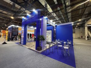 Estande da Água de Cheiro em uma feira de negócios, com estrutura azul, expositores de produtos e mesas para atendimento.