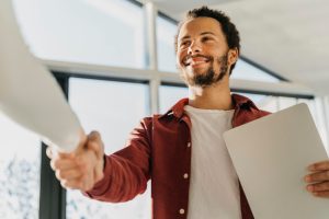 Homem sorridente aperta a mão de outra pessoa, simbolizando o fechamento de um acordo ou parceria, em ambiente corporativo.