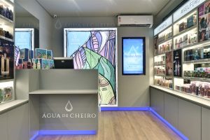 Interior de loja Água de Cheiro com balcão de atendimento, prateleiras iluminadas com perfumes e cosméticos e painel digital de divulgação.
