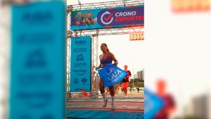 Mulher corredora cruza a linha de chegada de uma corrida de rua, segurando uma toalha azul com a marca “Água de Cheiro”, sob o pórtico da Crono Esportes.