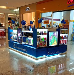 Quiosque da marca Água de Cheiro em shopping center, com prateleiras iluminadas exibindo perfumes, cosméticos e kits. Balões azuis decoram o espaço e telas digitais exibem campanhas da marca.