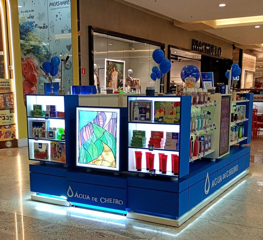 Quiosque da Água de Cheiro em um shopping, com exposição de perfumes e cosméticos iluminados em prateleiras azuis e decoração com balões azuis.