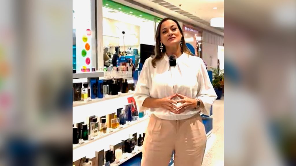 Mulher em pé dentro de uma loja de perfumes, falando para a câmera, com prateleiras cheias de fragrâncias ao fundo.