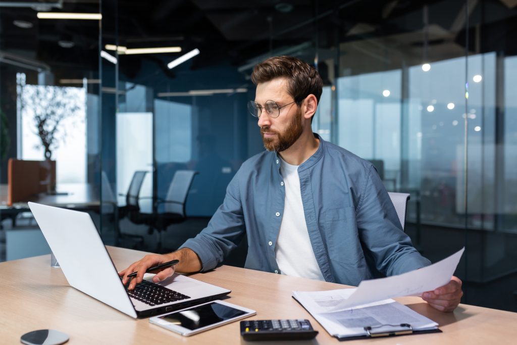 Homem usando óculos, concentrado enquanto analisa documentos e trabalha em um notebook, em um escritório moderno com divisórias de vidro — representando análise da COF no processo de abertura de uma franquia.