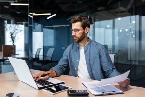 Homem usando óculos, concentrado enquanto analisa documentos e trabalha em um notebook, em um escritório moderno com divisórias de vidro — representando análise da COF no processo de abertura de uma franquia.